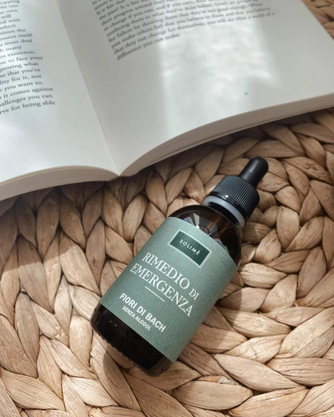 Steklenička Bachovih cvetnih kapljic Solimè »Rimedio di emergenza« brez alkohola, položena na naravno pleteno podlago ob odprti knjigi – simbol sprostitve, umirjenosti in ravnovesja.
